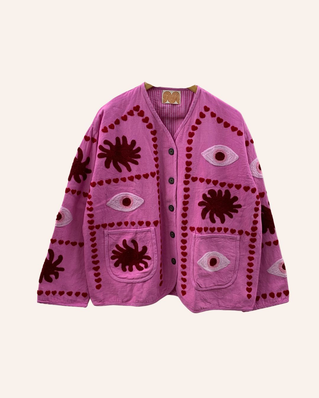 Front view of a pink hand-embroidered cotton jacket with eye and sunburst motifs, heart borders, and front pockets.