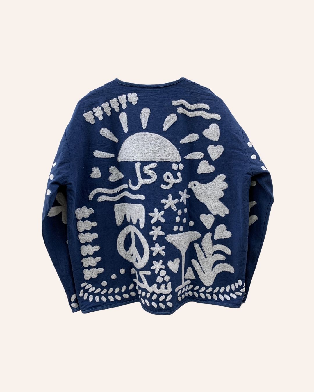 Back view of navy jacket with white embroidery showing the Arabic word for tawakkul, peace sign, rising sun, hearts, and nature-inspired symbols.