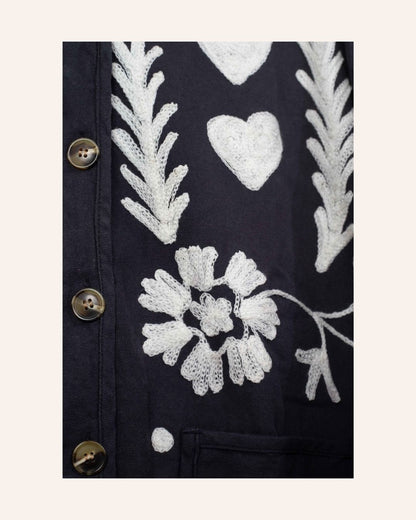 Hand-embroidered navy cotton jacket with white hearts, florals, and JOY motif on the back, inspired by playful folk-art storytelling.